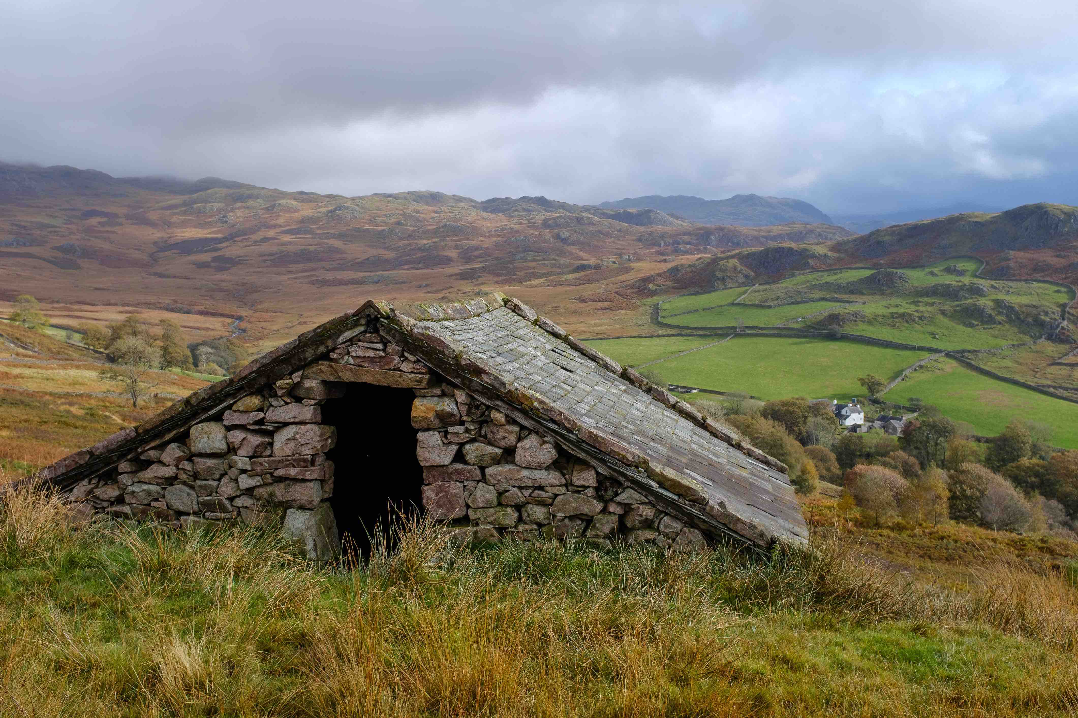 a stone hut in the foreground with green and orange meadows in the distance with hills and dry stone walls